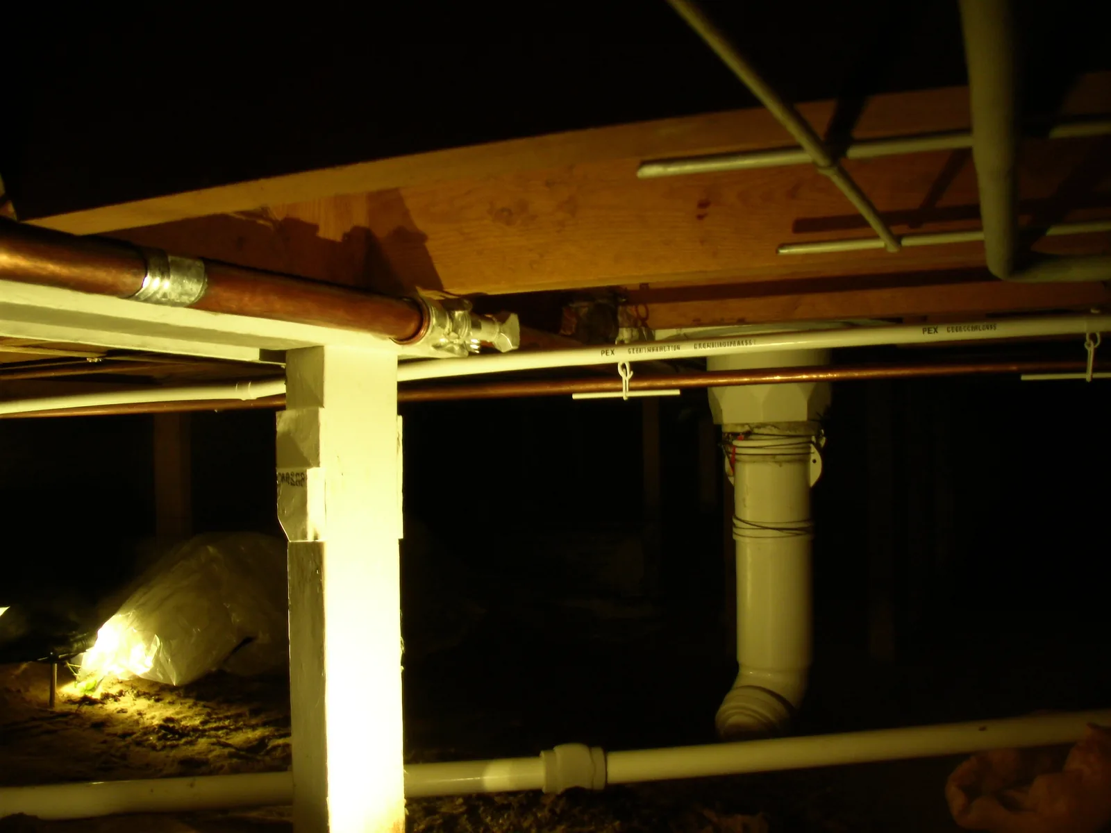 Copper and PEX water lines strapped to floor joists in a crawlspace, lit by a single work light, with a PVC drain line and cleanout below.