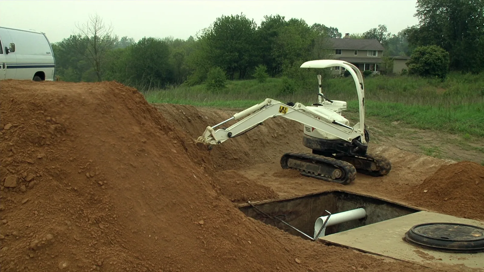 A concrete septic tank uncovered in a rural yard, with a mini excavator on the spoil pile and a service van parked at the edge of the field.